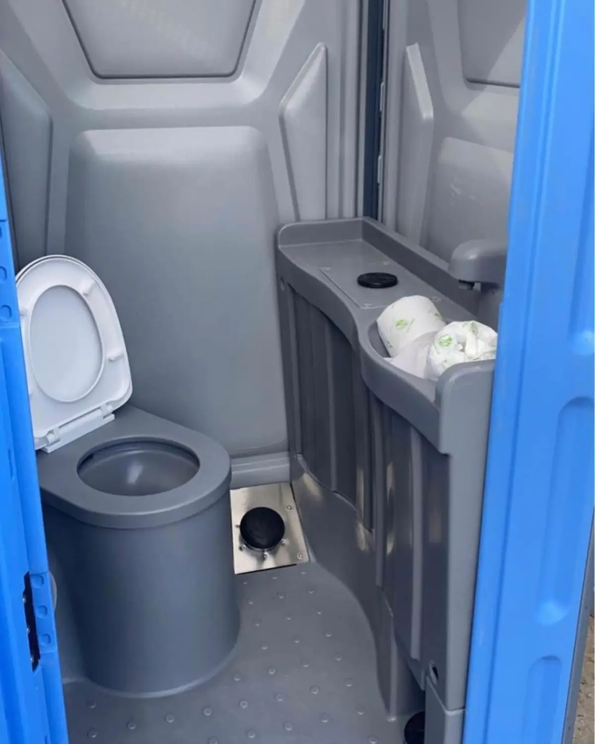 Inside a flushing portable toilet with a white seat, paper rolls and a sink tray