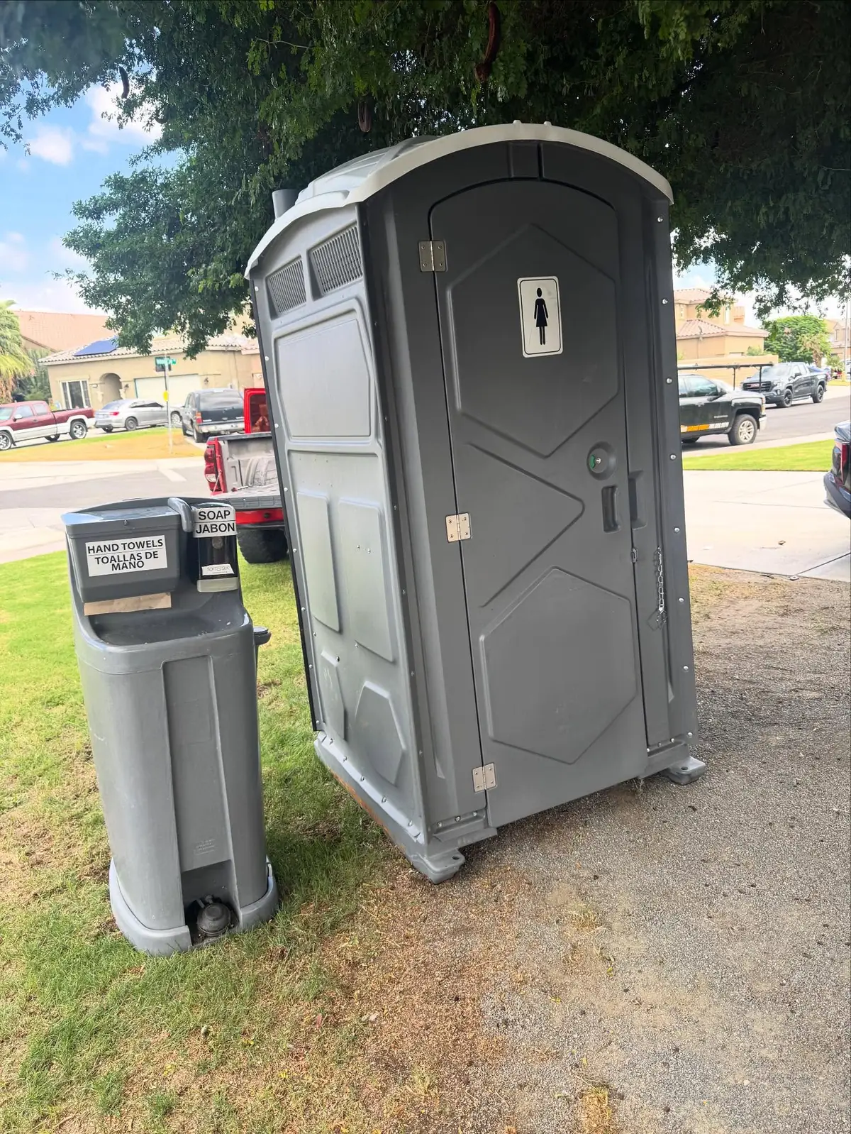Freestanding gray hand washing station beside a gray portable toilet on a lawn